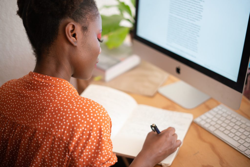 mulher negra escrevendo em seu caderno, em frente a um computador de mesa.